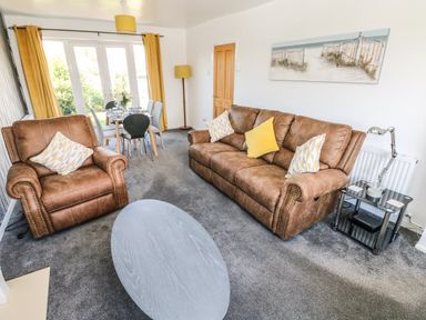 A living room with brown sofas a grey oval coffee table dining table with six chairs yellow curtains and a painting on the wall at 53 St Brides View in Roch