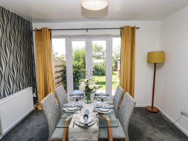 A dining room with glass table and six chairs set for a meal with yellow curtains and a floor lamp at 53 St Brides View in Roch