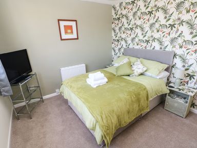 A bedroom with a bed covered with green bedding and pillows a nightstand with a lamp a television on a metal stand and a framed picture on the wall at 53 St Brides View in Roch
