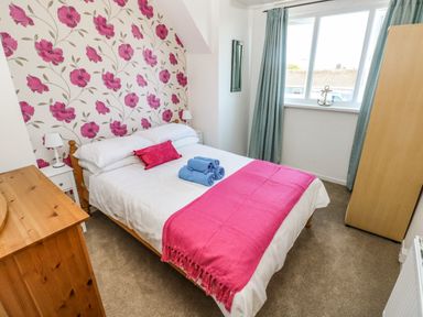 A bedroom with a bed covered with white and pink bedding beside wooden furniture and a window with blue curtains at 53 St. Brides View in Roch
