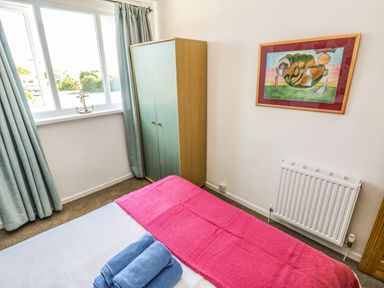 A bedroom with a bed covered by a pink blanket and blue towels folded on top a green wardrobe by the window with blue curtains and a framed abstract painting on the wall at 53 St Brides View in Roch
