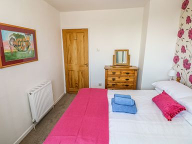 A bedroom with a bed covered in white and pink bedding with blue towels on it a wooden dresser with a mirror a wooden door and a painting on the wall at 53 St Brides View in Roch