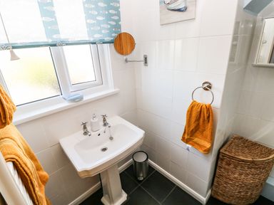 A bathroom with a white pedestal sink yellow towels a wicker laundry basket a round wall mirror and a window with a fish patterned blind at 53 St Brides View in Roch