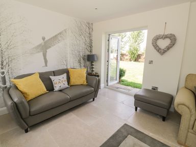 A living room with a gray sofa with yellow pillows a small table with a lamp an ottoman and an open door to a garden at Garden Cottage in Upton Upon Severn