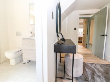 A hallway with a black console table and round stool next to a bathroom with a toilet and vanity at 12 Manor Farm Close in Kingham