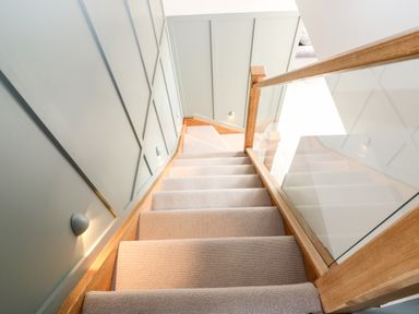 A carpeted staircase with wooden handrail and glass panel on one side and grey paneled wall on the other at 12 Manor Farm Close in Kingham