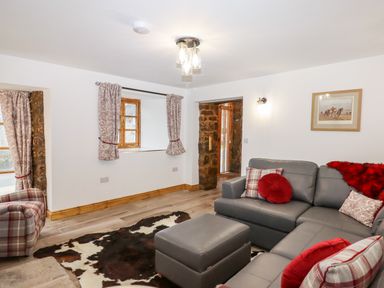 A living room with a gray sectional sofa red and plaid cushions a plaid armchair cowhide rug wooden floor and white walls at Bagtor Granary in Ilsington