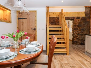A dining area with a wooden table set with plates bowls glasses and a vase with red tulips next to wooden stairs and a stone wall at Bagtor Granary Ilsington