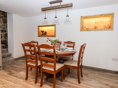 A dining room with a wooden round table set with plates glasses and cups six wooden chairs a flower vase on the table two built in wall shelves with lighting and decorative items wooden floor Bagtor Granary Ilsington