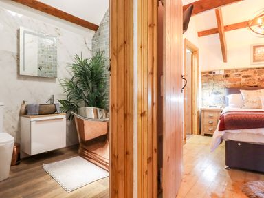 A bathroom with a metal bathtub, sink, mirror, and plant next to a bedroom with a bed, nightstand, and exposed wooden beams at Bagtor Granary in Ilsington