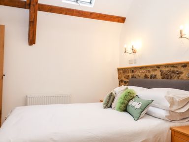 A bedroom with a white bed and decorative pillows stone wall section wooden beams and wall lights at Bagtor Granary in Ilsington