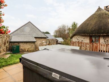 An outdoor area with a covered hot tub, stone buildings with slate and thatched roofs, and a wooden fence at Bagtor Granary in Ilsington