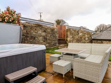An outdoor patio area with a hot tub and white wicker furniture with table and stools by a stone wall with a wooden gate at Bagtor Granary in Ilsington