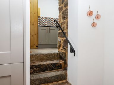 Stone steps leading up to a kitchen area with gray cabinets and patterned tile backsplash with copper pans hanging on a white wall at Bagtor Granary in Ilsington