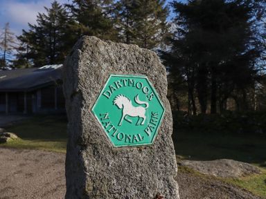 A stone pillar with a Dartmoor National Park sign in a wooded area with a building in the background