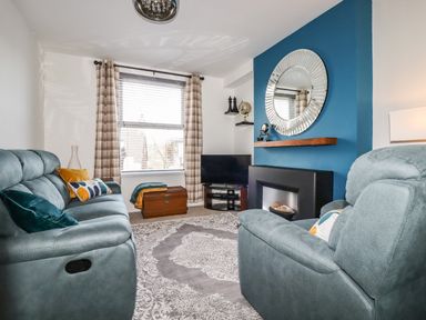 A living room with teal sofas cushions a TV on a corner stand a fireplace with a round mirror above and checked curtains at Penmar in Looe