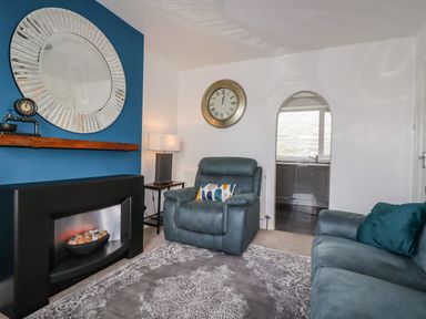 A living room with a grey armchair and sofa a wall clock and a mirror above a fireplace at Penmar in Looe
