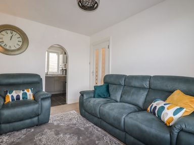A living room with a blue sofa and armchair with cushions a wall clock and an arched doorway to a kitchen at Penmar in Looe