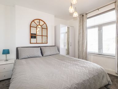 A bedroom with a double bed covered in gray bedding a white nightstand with a blue lamp a white wardrobe and a window with blinds and curtains at Penmar in Looe