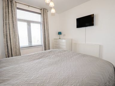 A bedroom with a bed covered in a quilted bedspread a window with blinds and curtains a white dresser with a lamp and a wall-mounted TV at Penmar in Looe