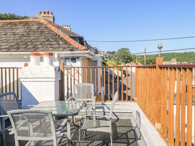 A rooftop patio with a glass table and metal chairs with wooden railing and rooftops in the background at Penmar in Looe