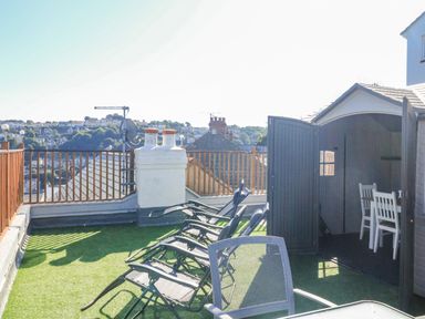 A rooftop terrace with lounge chairs a shed with an open door showing chairs and a table and a railing overlooking houses and trees at Penmar in Looe