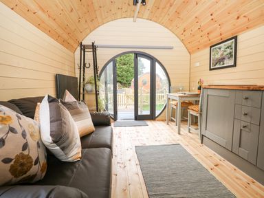 An interior view of a wooden pod with a black sofa with pillows on the left side a dining table with chairs on the right and a circular glass door at the far end at Filbert Pod in Arundel West Sussex