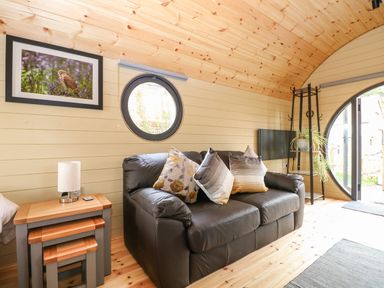A living room with a black leather sofa with cushions nest of tables a lamp plant a TV and round windows at Filbert Pod in Arundel West Sussex