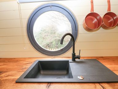 A kitchen sink below a round window with three orange pans hanging on the wall at Filbert Pod in Arundel West Sussex