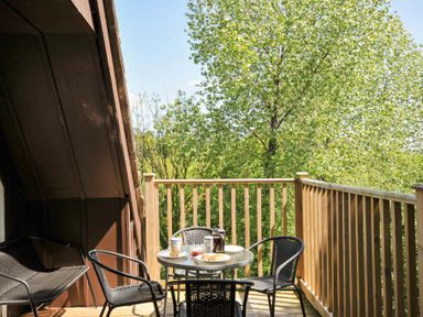 A balcony with a glass table and four black chairs surrounded by a wooden railing and trees at 60 Valley Lodge in Callington near Gunnislake