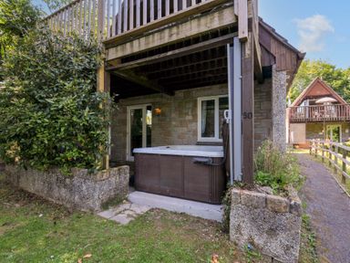 An outdoor hot tub under a wooden balcony next to a stone wall house at 60 Valley Lodge in Callington near Gunnislake