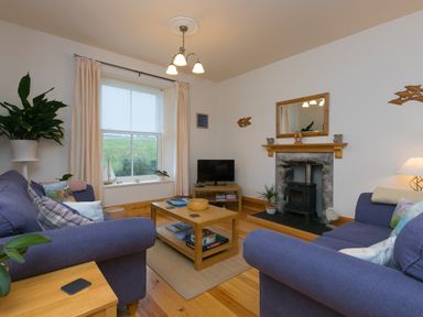 A living room with two blue sofas a wooden coffee table a fireplace a television and a window with curtains at Chynance in Porthcurno