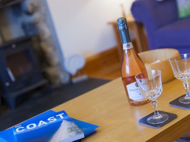 A wooden table with a bottle of Pinot Rose wine two wine glasses blue books and part of a purple couch in a living room at Chynance in Porthcurno