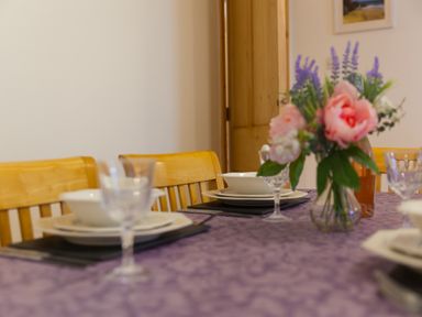 A dining table with a purple tablecloth set with plates bowls wine glasses and a vase of flowers at Chynance in Porthcurno