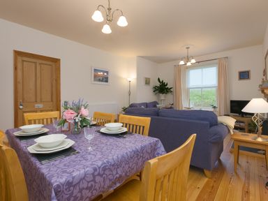 A dining area with a table set with plates and glasses next to a living room with blue sofas and a television at Chynance in Porthcurno