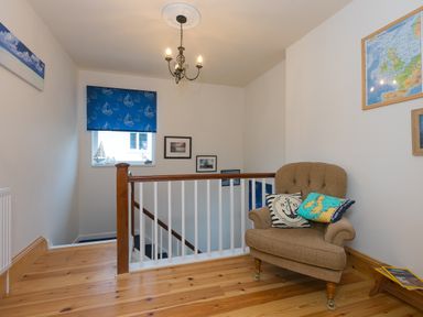 A stair landing with a brown armchair and decorative cushions a window with a blue sailboat blind framed pictures on the walls and wooden flooring at Chynance in Porthcurno