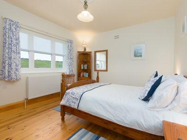 A bedroom with wooden bed frame blue and white bedding wooden bookcase and window with blue floral curtains at Chynance in Porthcurno