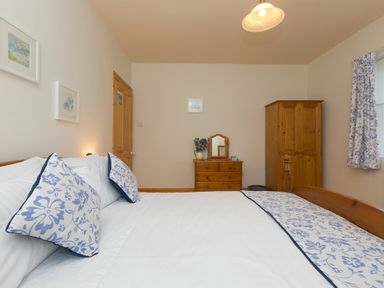 A bedroom with a bed with blue and white floral pillows and bedspread a wooden wardrobe wooden dresser with mirror and a window with floral curtains at Chynance in Porthcurno