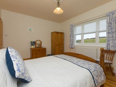 A bedroom with a wooden bed chest of drawers wardrobe chair and a window with floral curtains at Chynance in Porthcurno