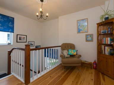 A landing area with a beige armchair and wooden bookshelf beside a staircase with wooden handrails and blue window blinds at Chynance in Porthcurno