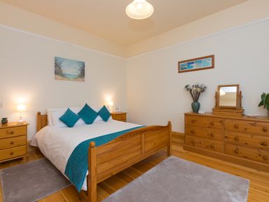 A bedroom with a wooden bed and blue cushions two bedside tables with lamps and a wooden dresser with a mirror at Chynance in Porthcurno