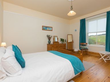 A bedroom with a wooden bed a dresser with mirror a standing mirror a wooden chair and a window with blue curtains at Chynance in Porthcurno