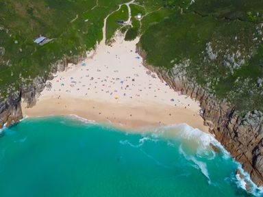 An aerial view of a sandy beach with people and umbrellas surrounded by green cliffs and turquoise ocean at Chynance in Porthcurno