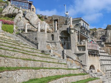 An outdoor stone amphitheater with tiered seating and a small building with lights and railings at Chynance in Porthcurno
