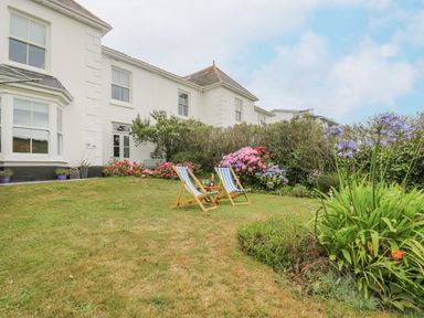 A garden with two wooden deck chairs and a small table in front of a white house with flowers and shrubs at Chynance in Porthcurno