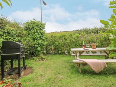 A garden with a barbecue grill and a wooden picnic table with a pink cloth and drinks at Chynance in Porthcurno