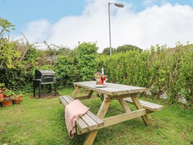 A wooden picnic table with wine and glasses next to a barbecue grill in a garden at Chynance in Porthcurno