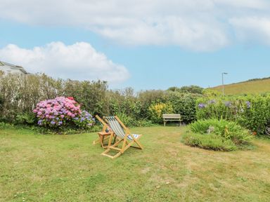 A garden with two deck chairs and a small wooden table surrounded by flower bushes and a wooden bench in the background at Chynance in Porthcurno