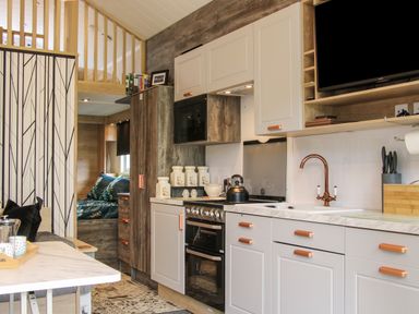 A compact kitchen with white cabinets and copper handles a black kettle on the stove and a television above the counter at Jacobs Meadow Pod 2 Minsterley near Chirbury
