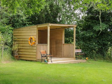 A wooden shed with an open front and two benches on a grassy area surrounded by trees at Jacobs Meadow Pod 2 Minsterley near Chirbury